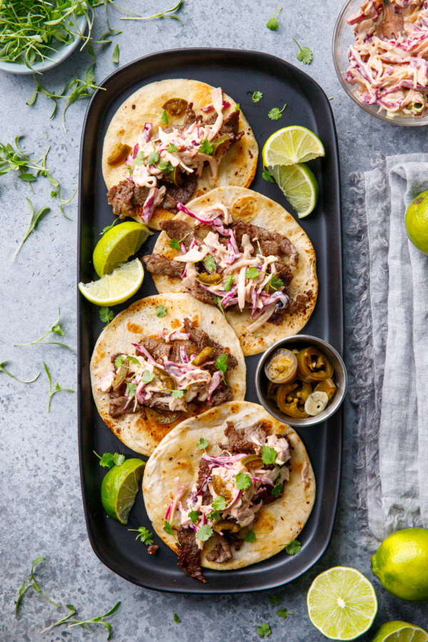 Overhead rectangular black plate with four street tacos with browned tortillas, slices of lime and bowls on the side