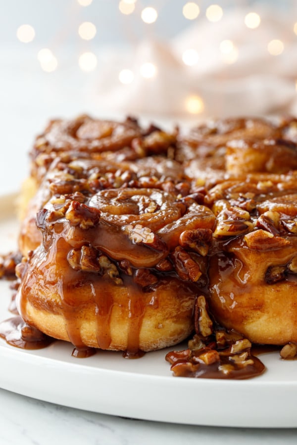 Gooey caramel dripping down the side of hot buttered rum sticky buns, with twinkle lights in the background