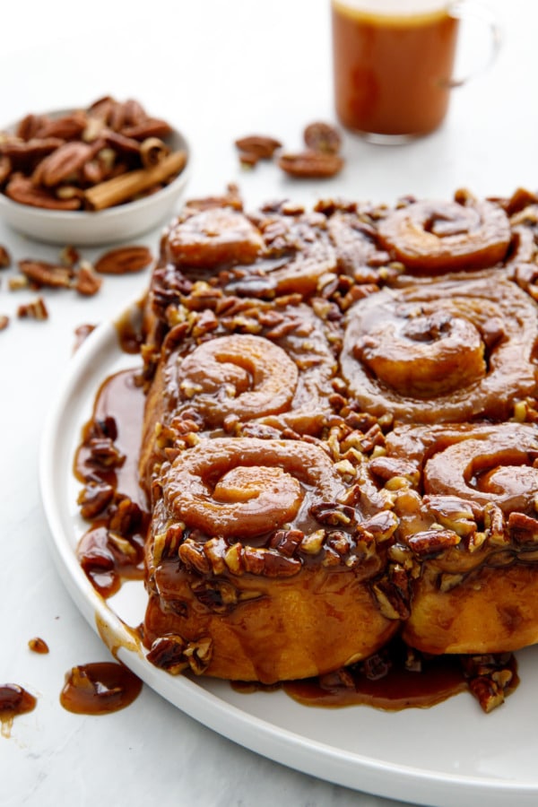 Platter of gooey sticky buns with dish of pecans and a cup of hot buttered rum in the background