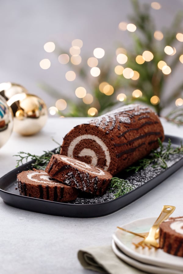 Sliced Chocolate Chestnut Christmas Cake Roll on a black rectangle platter, green and gold christmas decor in the background