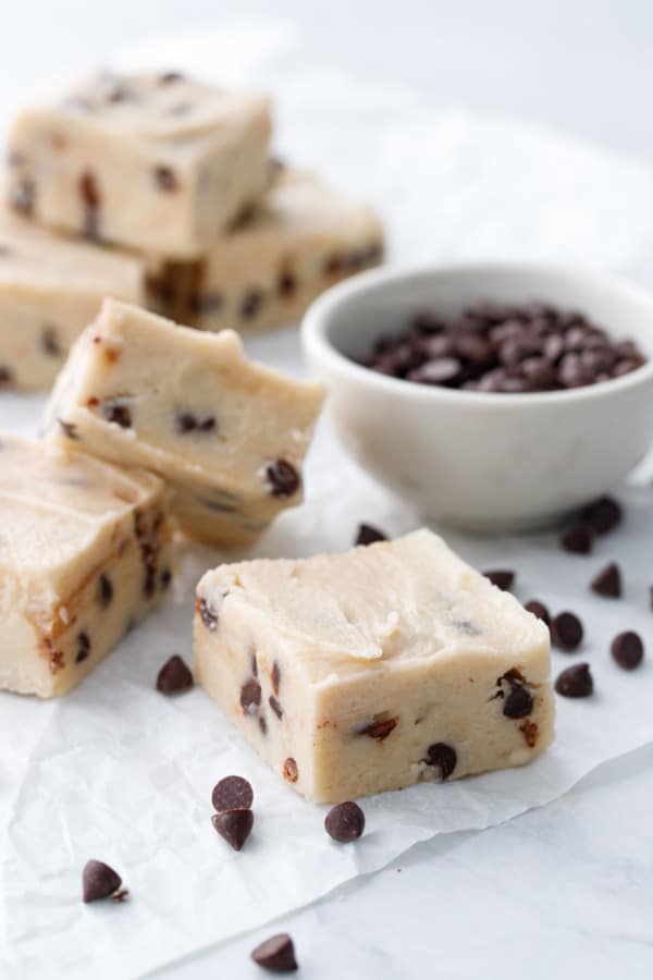 Cut pieces of Chocolate Chip Cookie Dough Fudge with a bowl of mini chocolate chips, on crinkled parchment