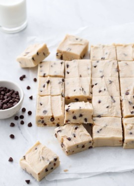 Cut pieces of Chocolate Chip Cookie Dough Fudge on parchment, with a glass of milk and bowl of mini chips