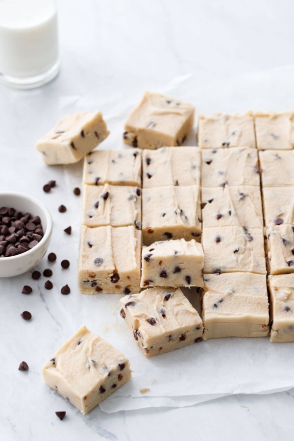 Cut pieces of Chocolate Chip Cookie Dough Fudge on parchment, with a glass of milk and bowl of mini chips