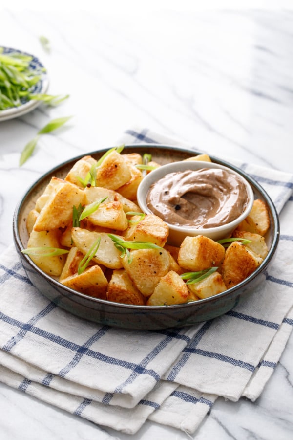 Shallow bowl filled with golden brown crispy potatoes, with a dish of black garlic aioli and sliced green onions