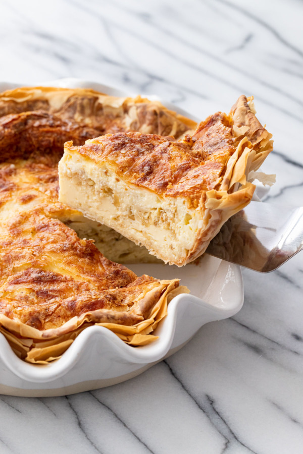 Lifting a slice of Cheese & Caramelized Onion Quiche out of a white ruffled pie dish