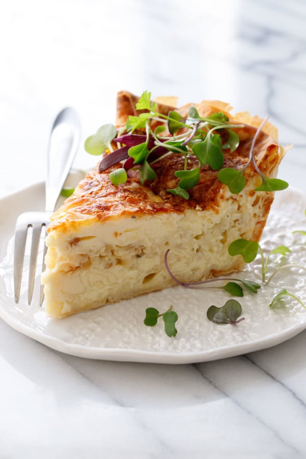 Slice of Cheese & Caramelized Onion Quiche topped with microgreens on a white ceramic plate and fork