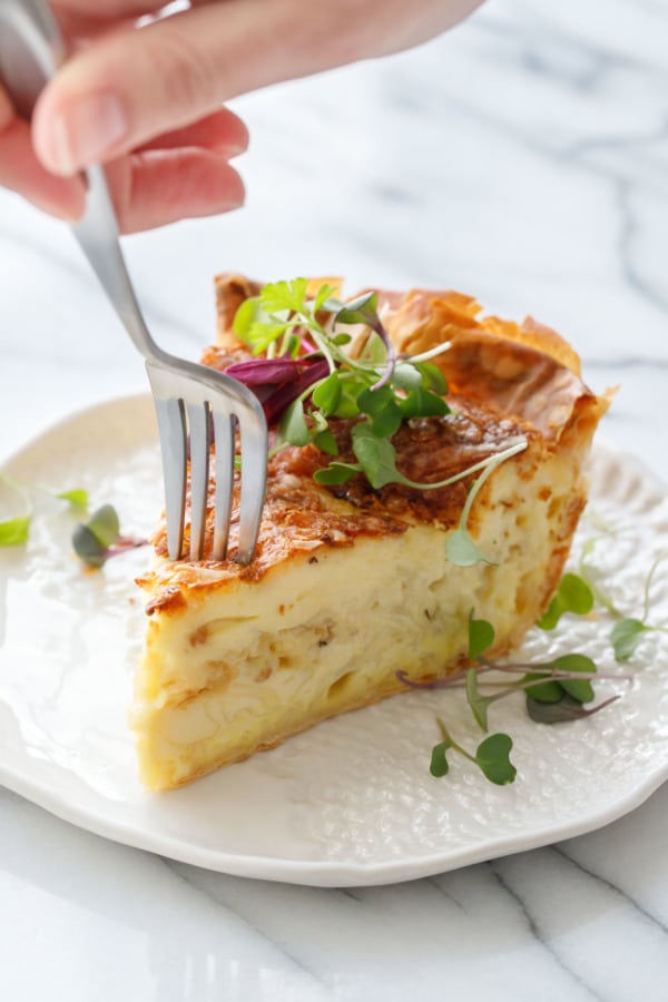 Fork in Slice of Cheese & Caramelized Onion Quiche topped with microgreens on a white ceramic plate and fork