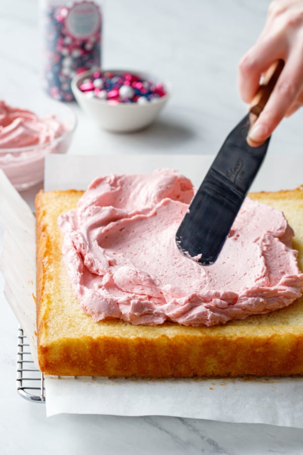 Offset spatula spreading pink frosting onto a yellow sheetcake