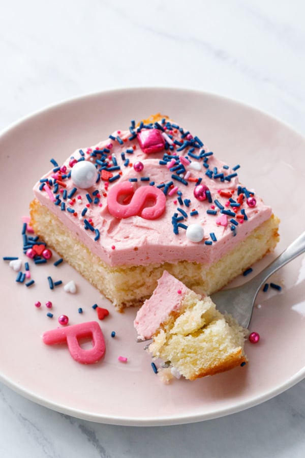 Slice and forkful of yellow cake with pink frosting on a pink plate with sprinkles