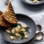 Two charcoal gray bowls with brothy white beans and wilted kale, with slices of grilled sourdough bread