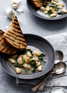 Two charcoal gray bowls with brothy white beans and wilted kale, with slices of grilled sourdough bread