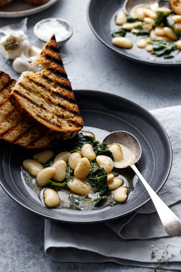 Ceramic bowl with brothy beans and kale, silver soup spoon and two slices of grilled bread.