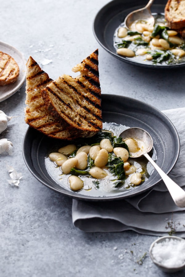 Two charcoal gray bowls with brothy white beans and wilted kale, with slices of grilled sourdough bread