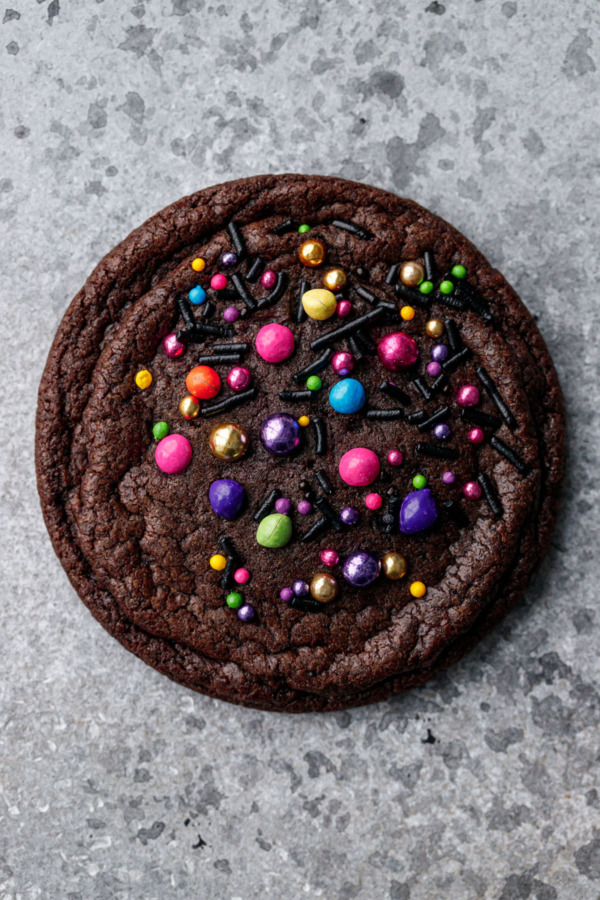 Single chocolate brownie cookie on a gray background with brightly colored rainbow and metallic sprinkles