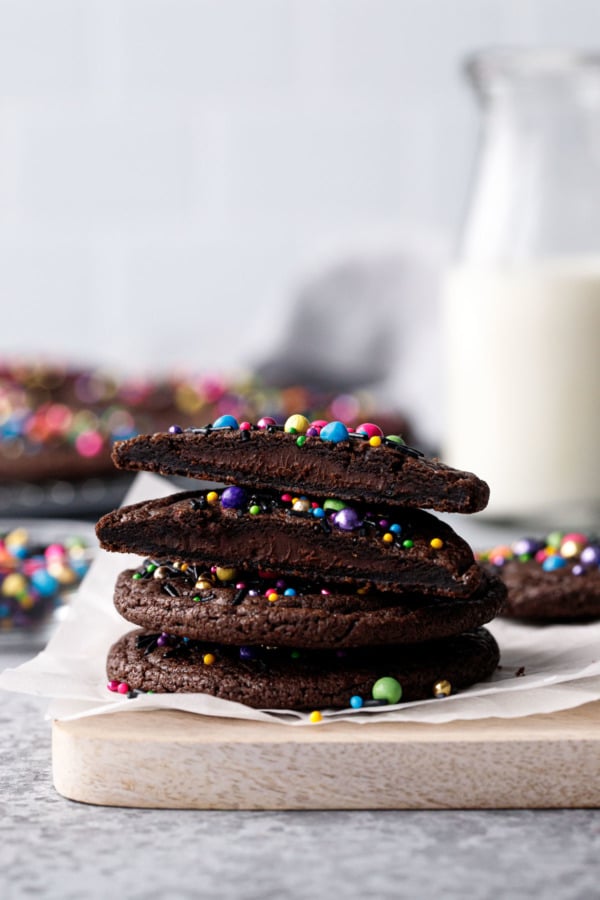 Stack of Cosmic Brownie Cookies, one cookie cut in half to show ganache filling; milk bottle and more cookies blurred in the background.