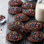 Cooling rack with Ganache-Stuffed Cosmic Brownie Cookies, one cut in half to show filling, and a small glass milk bottle