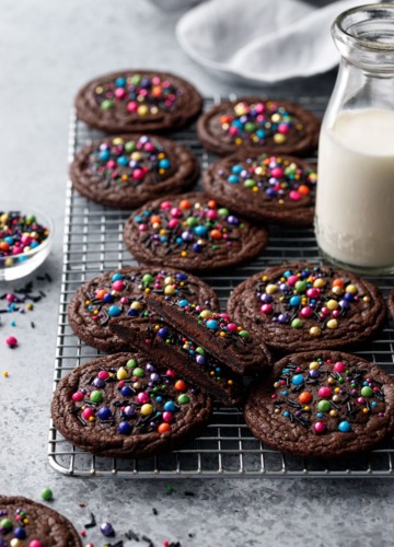 Cooling rack with Ganache-Stuffed Cosmic Brownie Cookies, one cut in half to show filling, and a small glass milk bottle