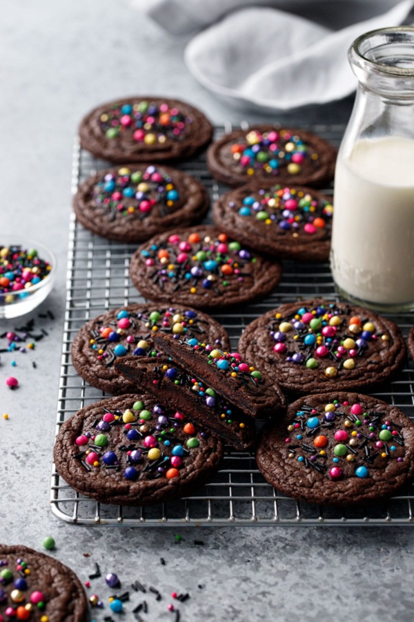 Cooling rack with Ganache-Stuffed Cosmic Brownie Cookies, one cut in half to show filling, and a small glass milk bottle