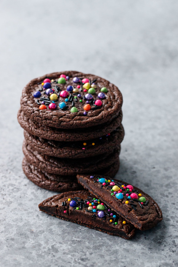 Stack of Ganache-Stuffed Cosmic Brownie Cookies on a gray background, one cookie cut in half to show the filling