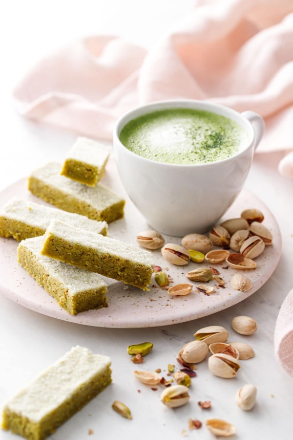 Fingers of Matcha Pistachio Shortbread on a ceramic plate with a mug of matcha and pistachios scattered around