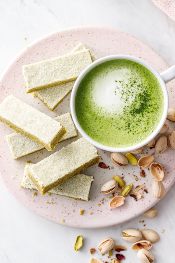 Overhead, fingers of Matcha Pistachio Shortbread on a pink ceramic plate with a white mug of matcha latte