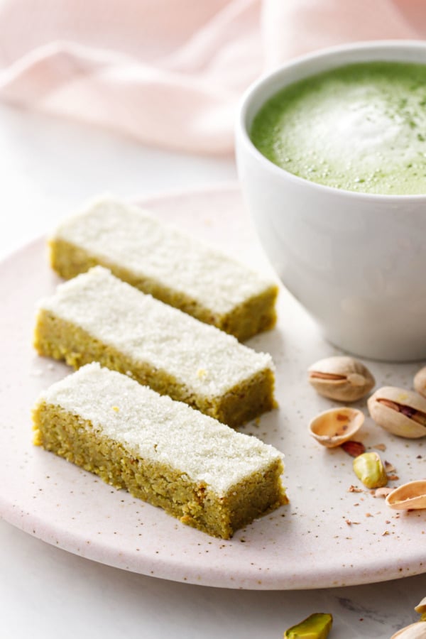 Three rectangles of Matcha Pistachio Shortbread on a pink ceramic plate with a matcha latte and pistachio on the side