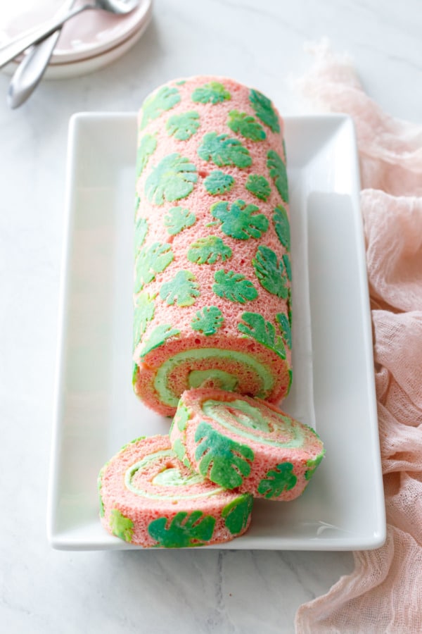 Pink cake roll with stenciled monstera leaf design, sliced to show the spiral of green pandan whipped cream filling