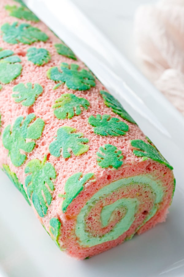 Pink cake roll with stenciled monstera leaf design, sliced to show the spiral of green pandan whipped cream filling