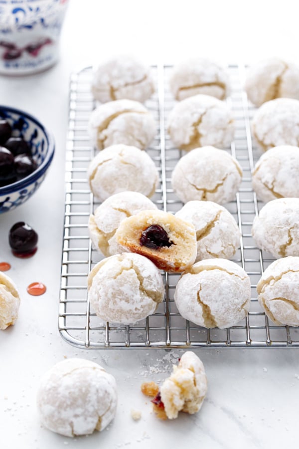 Wire rack with rows of amaretti cookies, one cookie cut in half to show the amarena cherry hidden inside.