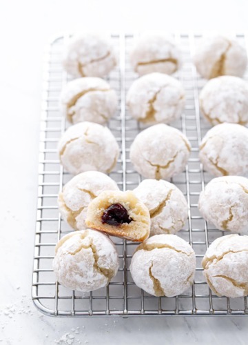 Wire rack with rows of amaretti cookies, one cookie cut in half to show the amarena cherry hidden inside.
