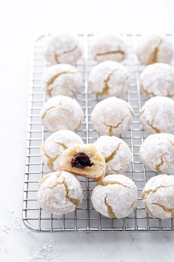 Wire rack with rows of amaretti cookies, one cookie cut in half to show the amarena cherry hidden inside.