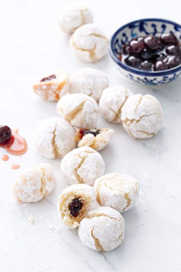 Scattered cookies, some cut in half to show the cherry filling, and a bowl of amarena cherries in the background.