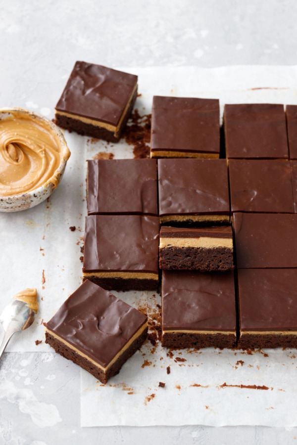 Sliced peanut butter ganache brownies on parchment, bowl of peanut butter on the side