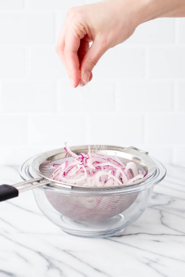 Sliced red onions in a sieve in a glass bowl, hand sprinkling with salt