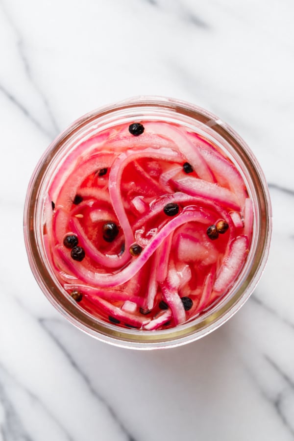 Overhead, open jar with pickled red onions and black peppercorns, on marble background