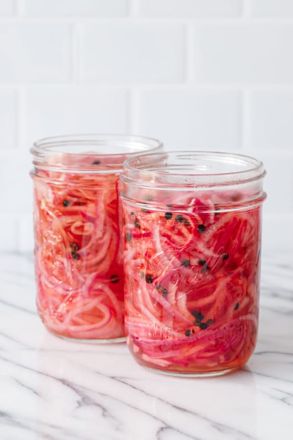 Two jars filled with sliced onions and vinegar brine, plus peppercorns dotted throughout.