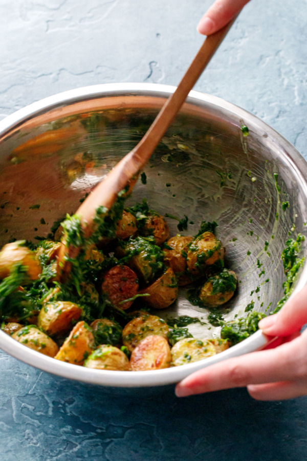 Metal bowl tossing roasted potatoes with arugula chimichurri