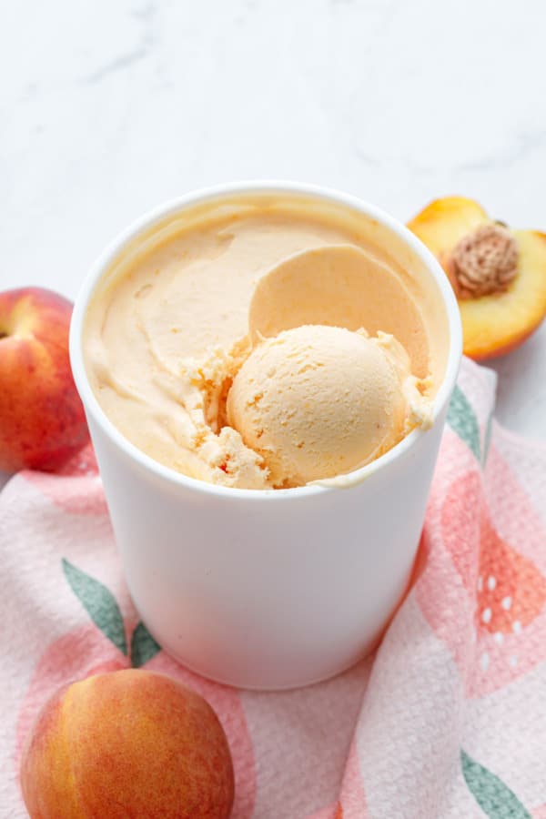 White container with peach ice cream and one scoop, sitting on a peach-graphic towel with fresh peaches