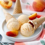 White plate with three scoops of peach ice cream, cones, peach towel and fresh peaches