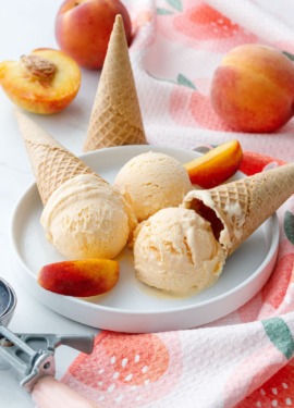 White plate with three scoops of peach ice cream, cones, peach towel and fresh peaches