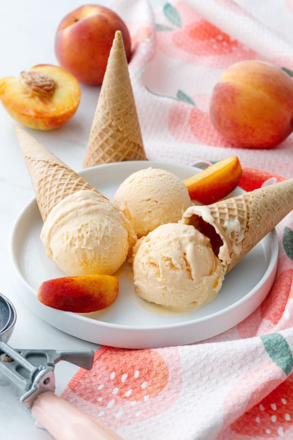 White plate with three scoops of peach ice cream, cones, peach towel and fresh peaches