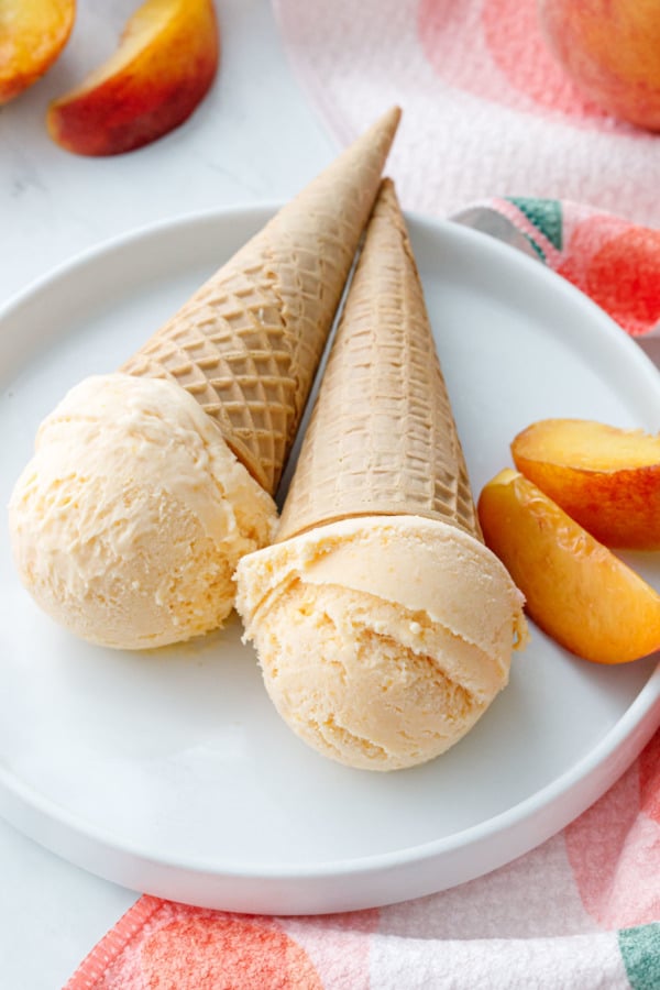 Two sugar cones with single scoops of fresh peach ice cream, peach slices on the side