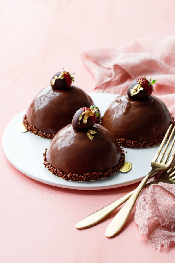 Three ganache-covered domes on a flat white plate, pink background, gold forks