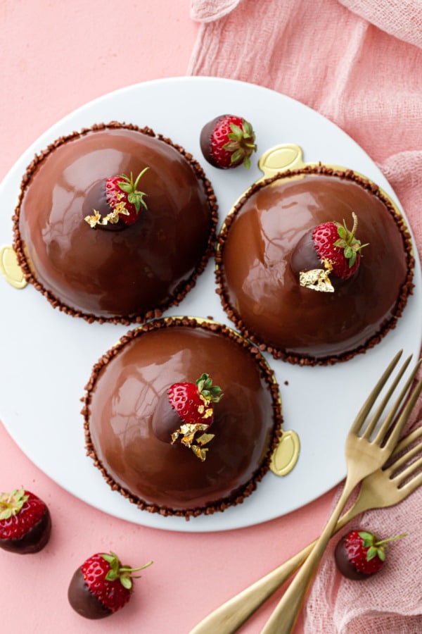 Overhead, three chocolate covered dome-shaped cakes on a white plate with gold forks