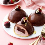 Dome-shaped strawberry mousse cakes, three on a white plate, one sliced to show the brownie inside