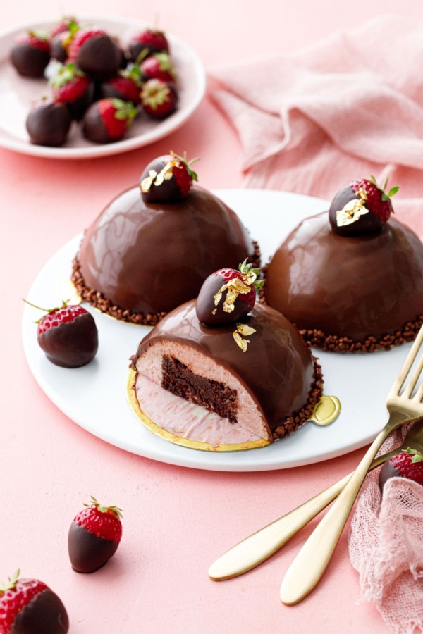 Dome-shaped strawberry mousse cakes, three on a white plate, one sliced to show the brownie inside