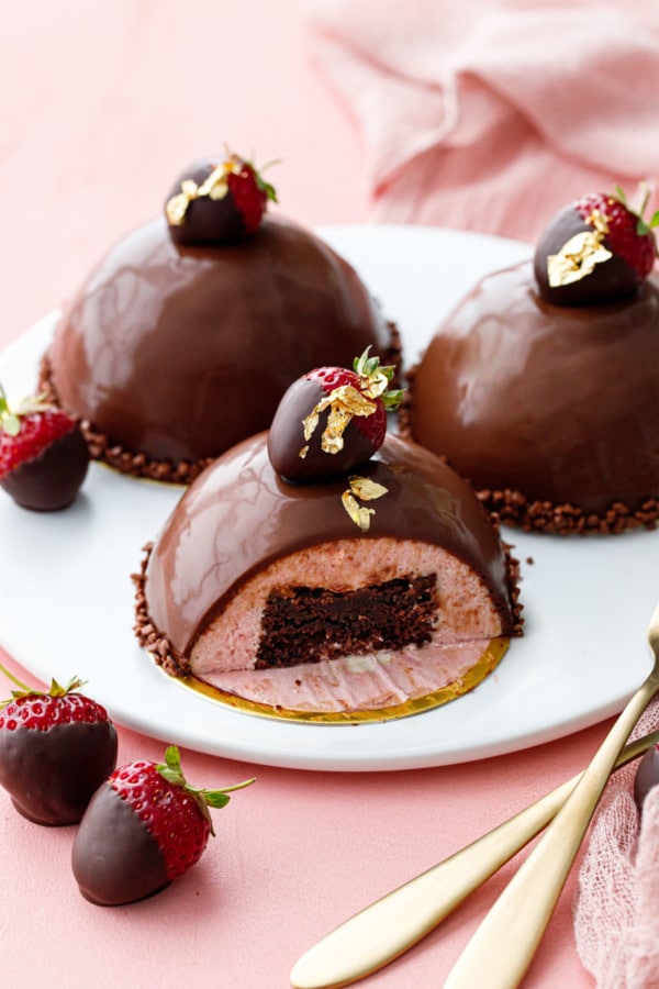 Strawberry mousse cakes covered in chocolate ganace, three on a white plate, one sliced to show the brownie inside