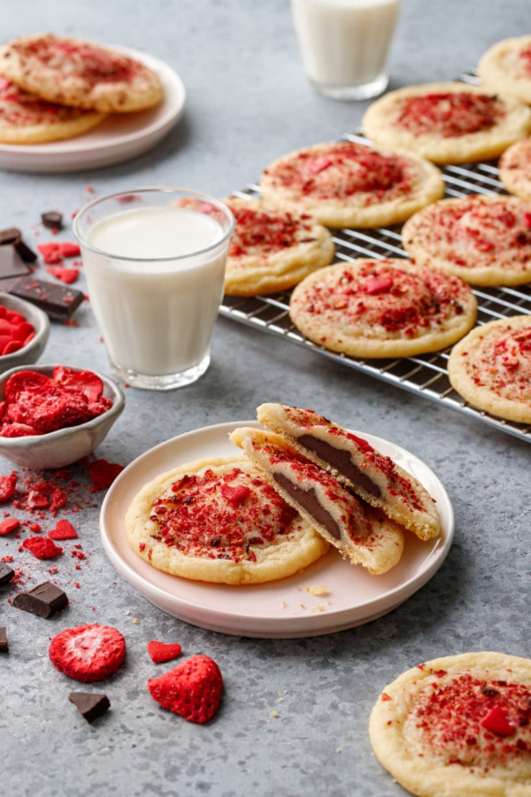 Plate with two cookies, one cut in half to show chocolate filling, with a rack of cookies, raw ingredients, and a glass of milk in the background.