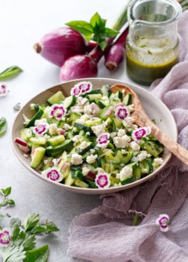 Cucumber salad garnished with edible flowers, in bowl with purple linen and red onions in background.