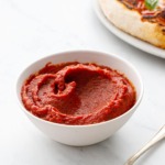 White bowl with homemade pizza sauce and spoon, homemade pizza on a plate in the background